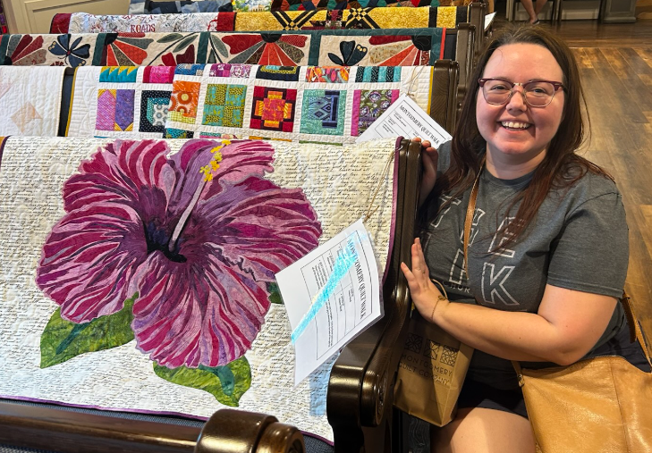 A smiling woman sits beside a colorful quilt displaying a large pink hibiscus flower, surrounded by various other quilts in a quilt show at a church. 